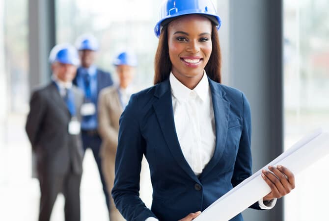 An architect in a hard hat holding drawings, colleagues reviewing plans behind her