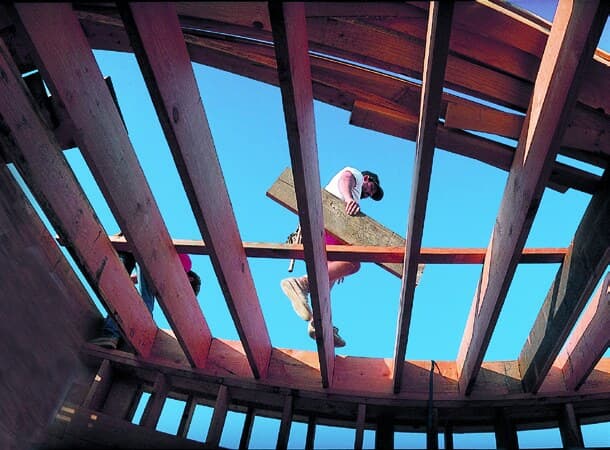 Looking up through open roof framing toward a worker and blue sky